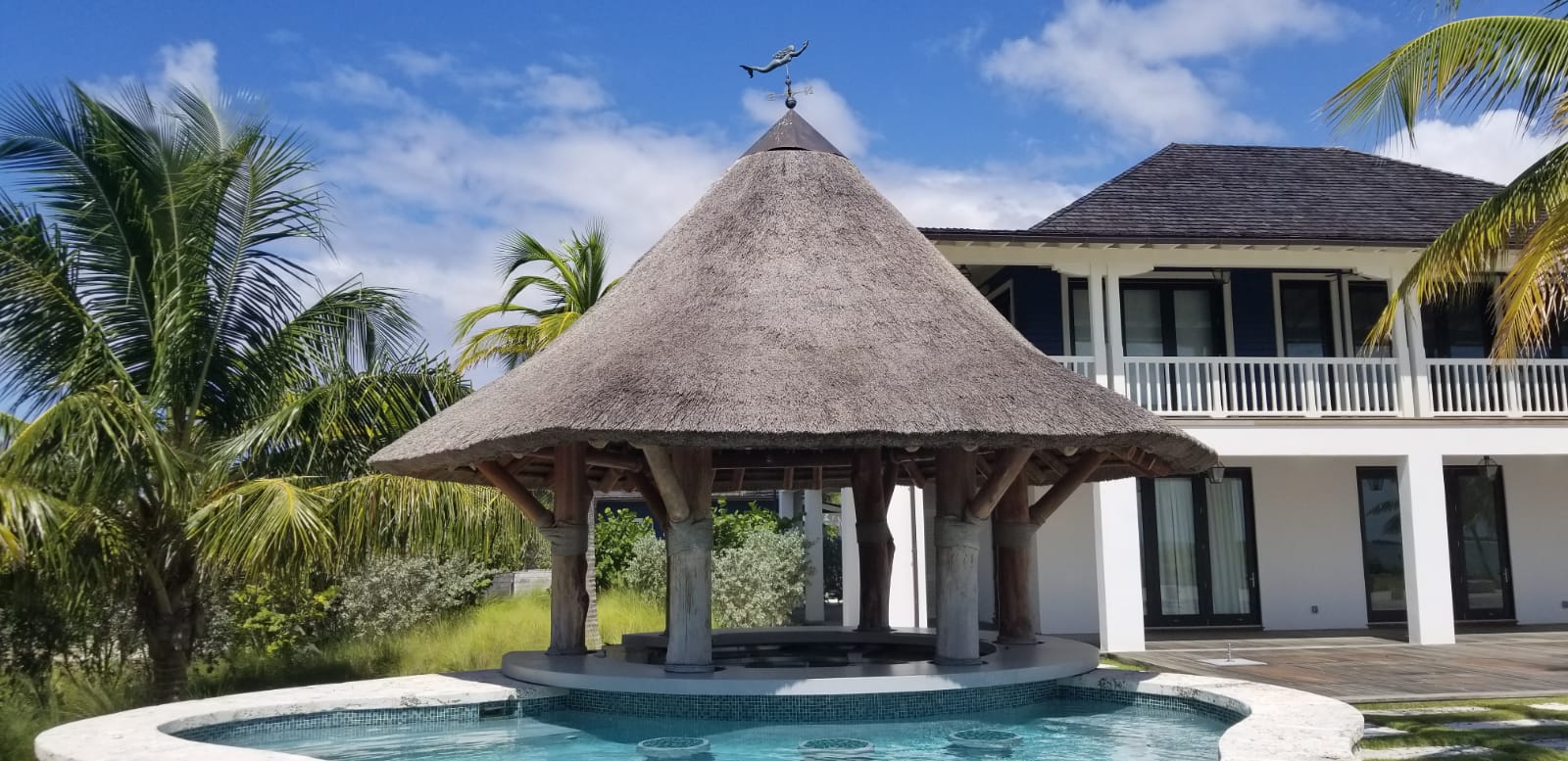 Tiki hut structure with thatched roof beside a pool, surrounded by palm trees and modern architecture, representing outdoor hospitality design by Tiki Hut Company.