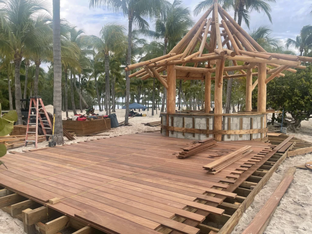 Tropical outdoor construction site featuring a wooden tiki hut structure with a stone base, surrounded by palm trees and sandy ground, showcasing custom landscaping and fencing solutions.