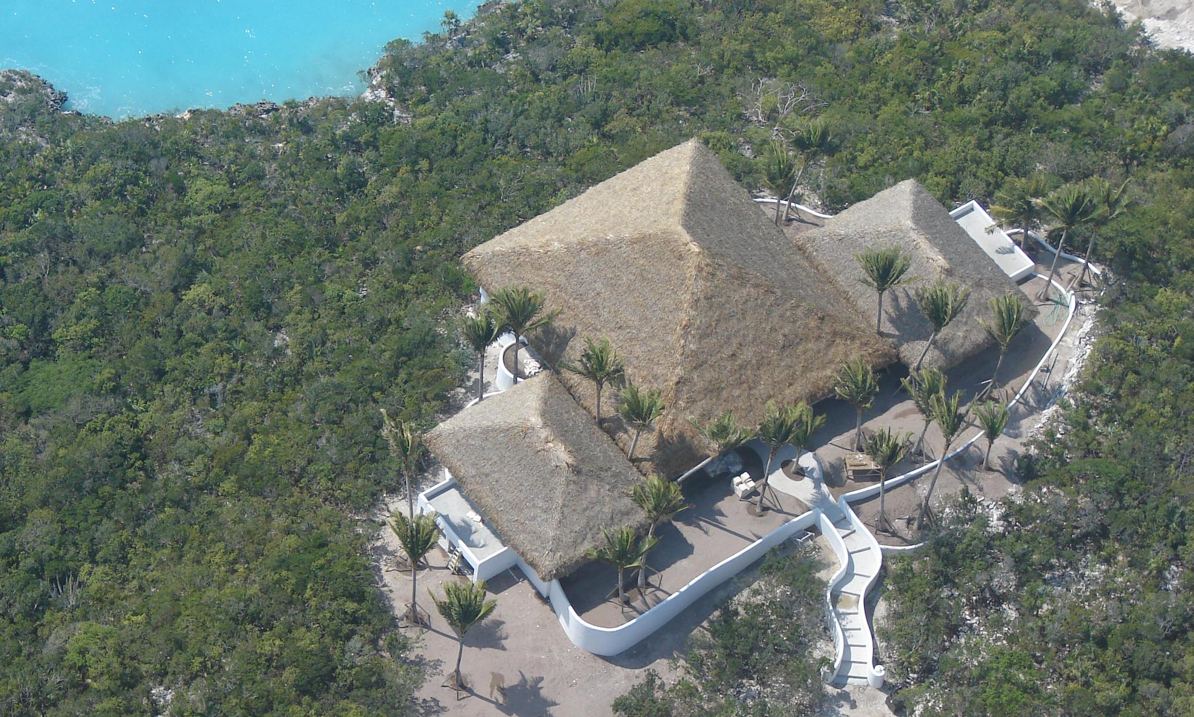 Aerial view of a Tiki Hut with thatched roofing surrounded by lush greenery and a turquoise water backdrop, emphasizing outdoor design and natural elements.