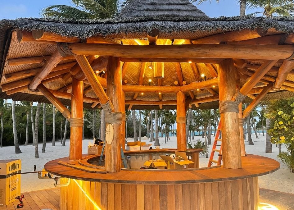 Tiki hut structure showcasing natural wood craftsmanship and tropical design, surrounded by palm trees and sandy beach, emphasizing outdoor luxury and landscaping services.