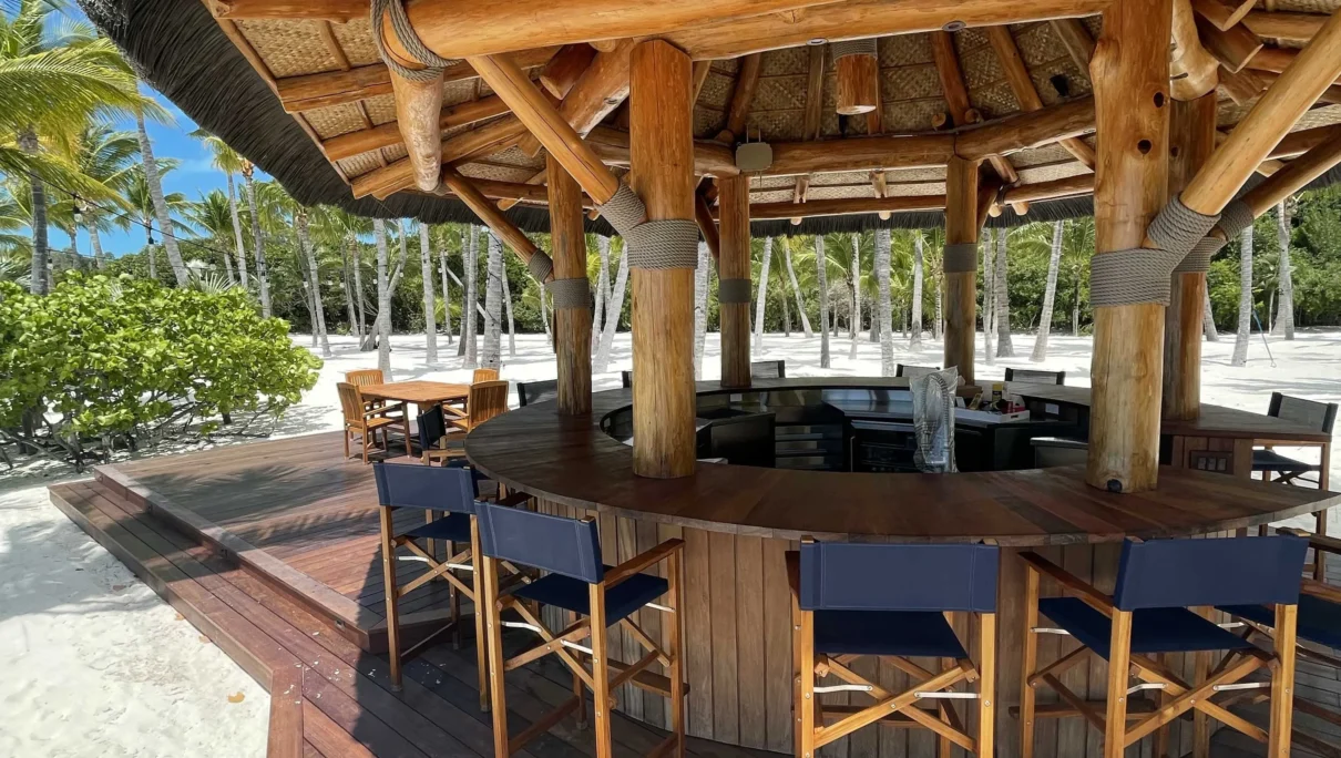 Tiki hut with a thatch roof and wooden bar seating, surrounded by palm trees and sandy beach, emphasizing sustainable outdoor living and tropical aesthetics.