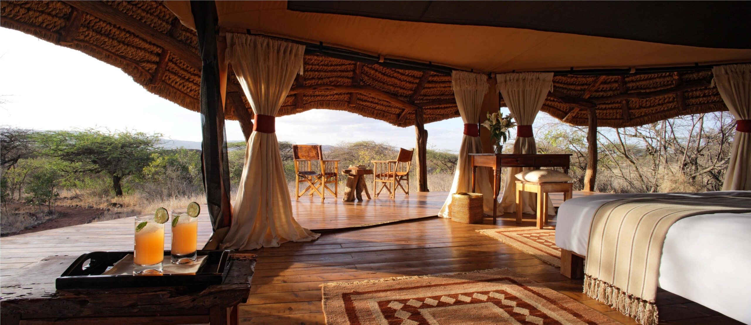 Luxurious tented accommodation featuring wooden flooring, a bed with decorative throw, and a scenic view of nature, including two refreshing drinks on a tray, emphasizing the Tiki Hut Company's expertise in creating immersive resort environments.