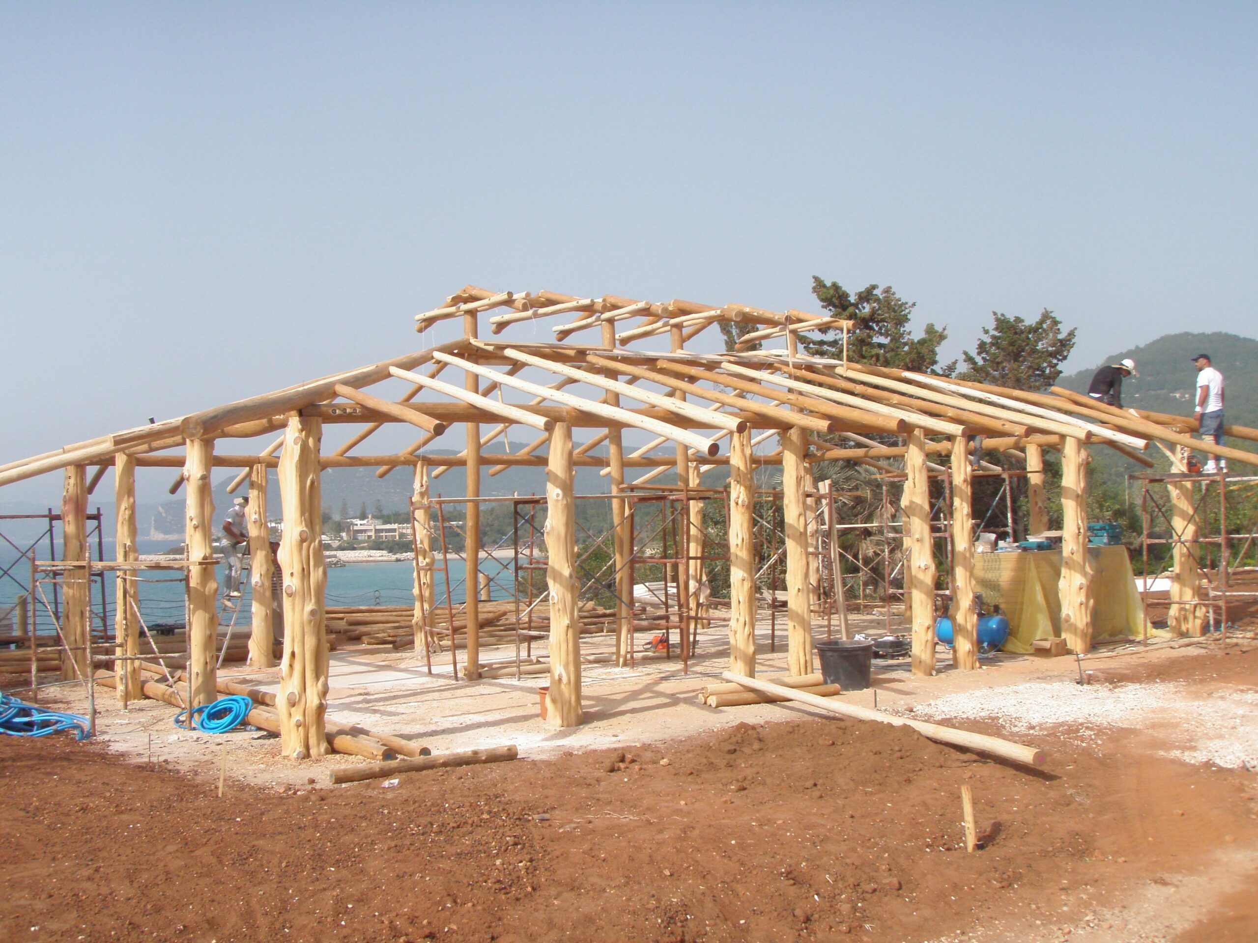 Tiki hut structure under construction with wooden beams, scaffolding, and workers, showcasing craftsmanship for residential and commercial tiki construction by The Tiki Hut Company.