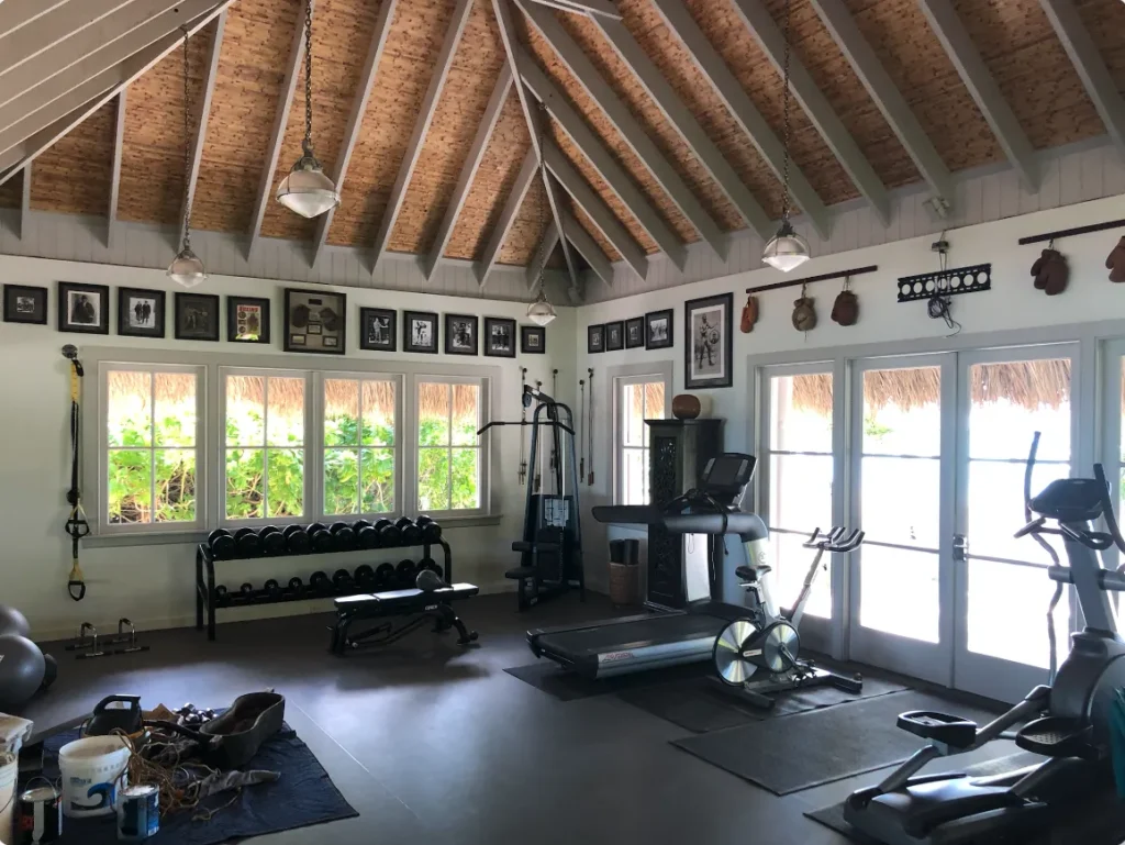 Interior of a fitness space featuring artificial thatch roofing, equipped with exercise machines, dumbbells, and framed photographs on the walls, highlighting eco-friendly design and aesthetic appeal.
